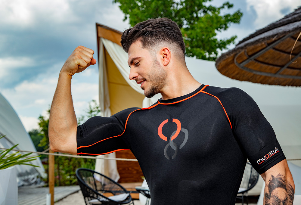 A man wearing a myostyle wireless ems training suit while flexing his biceps outdoors.